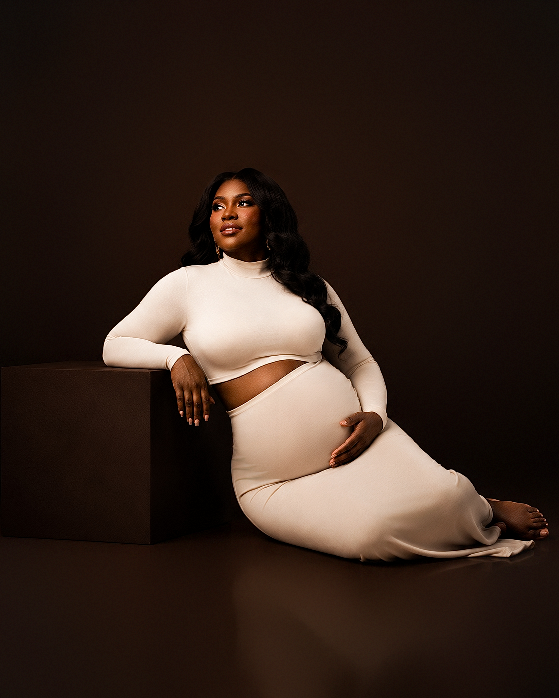 Expectant mother in a cream knit two-piece seated on a studio block with one hand on her belly, maternity portrait