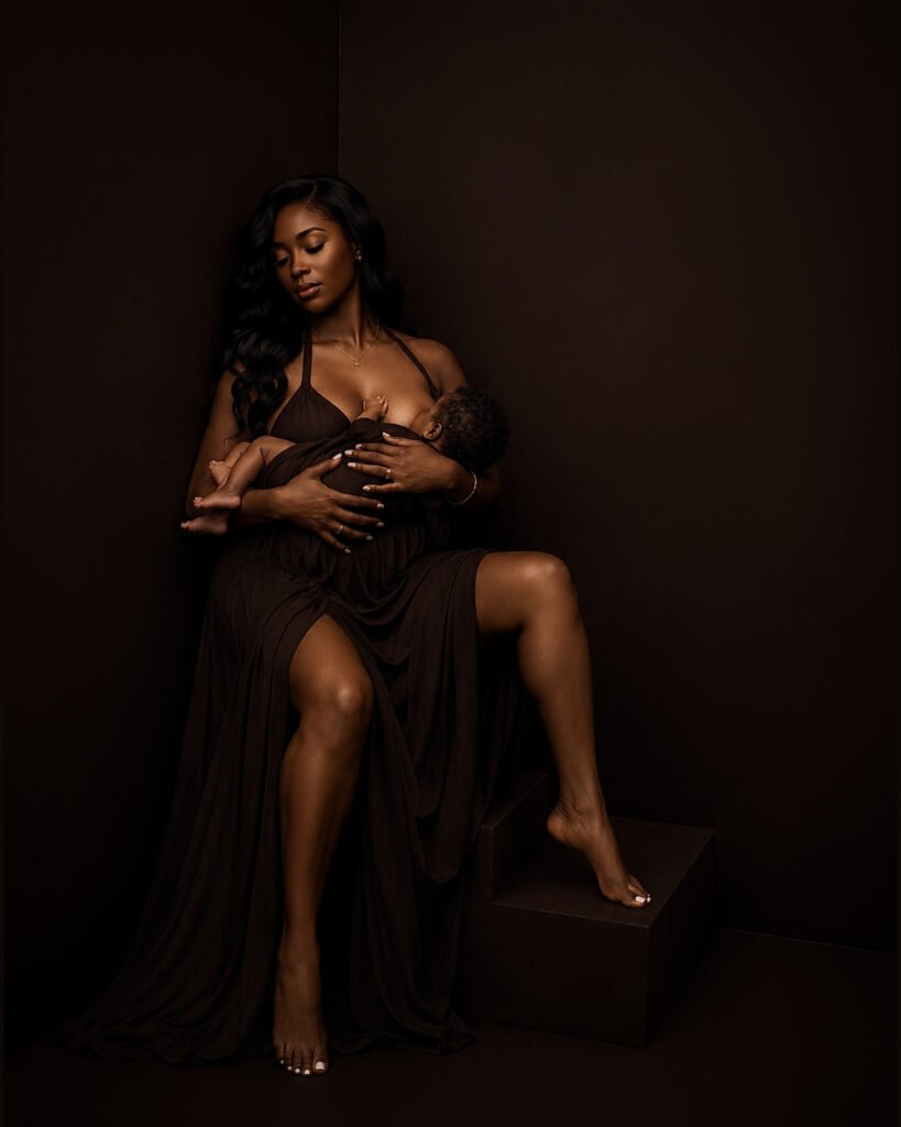 Mother breastfeeding her newborn while seated in a black gown in warm low light, newborn portrait