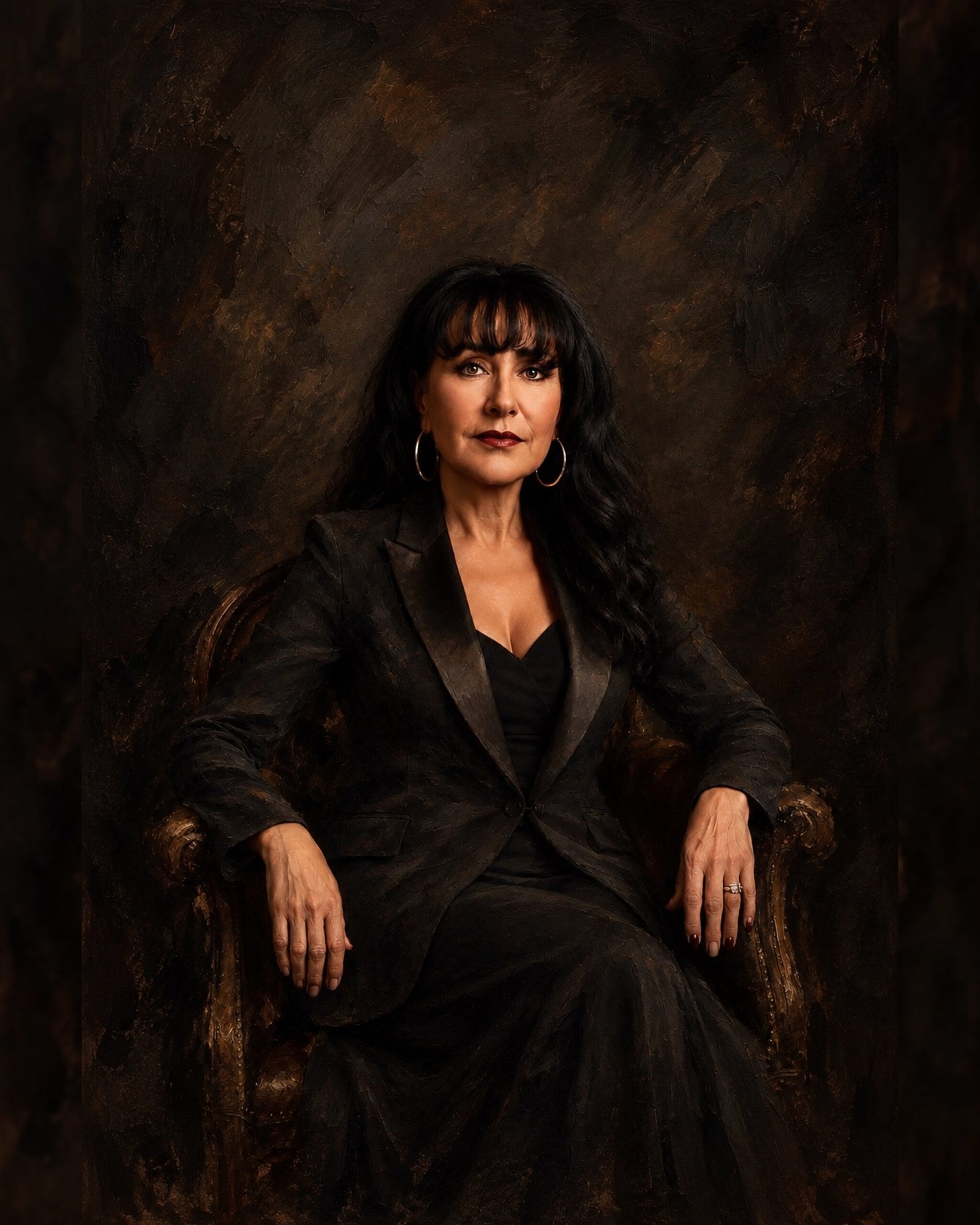 Woman in a black suit seated against a dark backdrop, looking directly at the camera in warm low light, women’s portrait in Hamilton