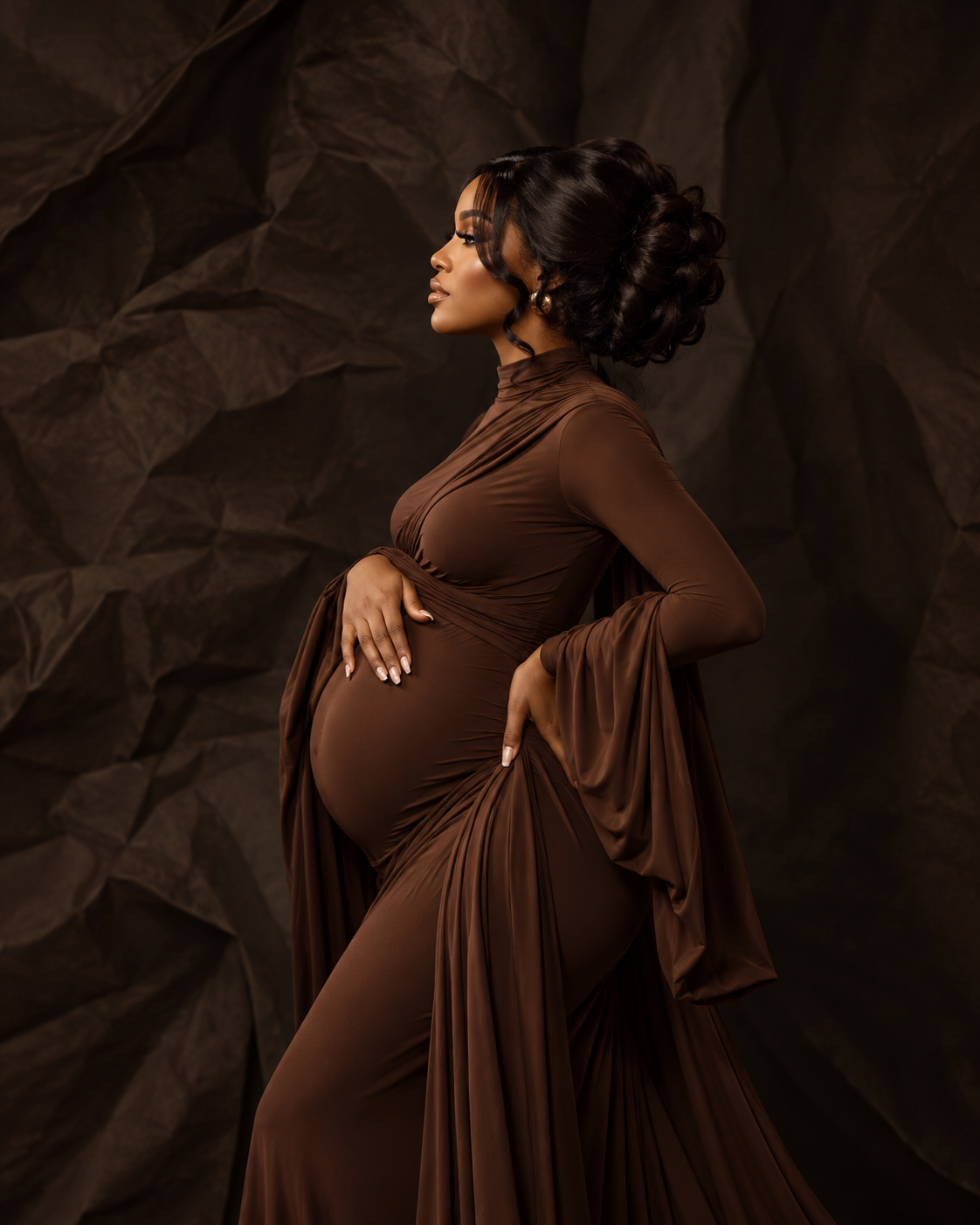 Expectant mother in brown fitted dress with one hand above her belly in warm low light, maternity portrait